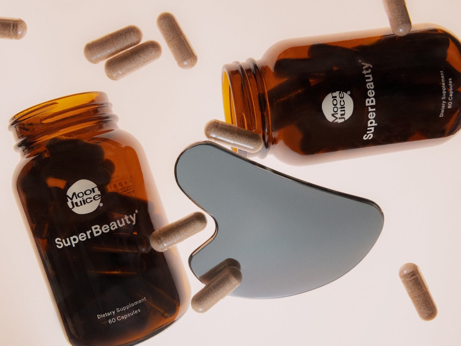 Two amber supplement bottles labeled Moon Juice SuperBeauty with several capsules scattered around and a dark gray applicator patch on a clean white background.