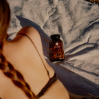 An amber supplement bottle labeled 'SuperHair' sits on a wrinkled light gray blanket, with a woman's braided hair and bare shoulder in the foreground under warm sunlight.