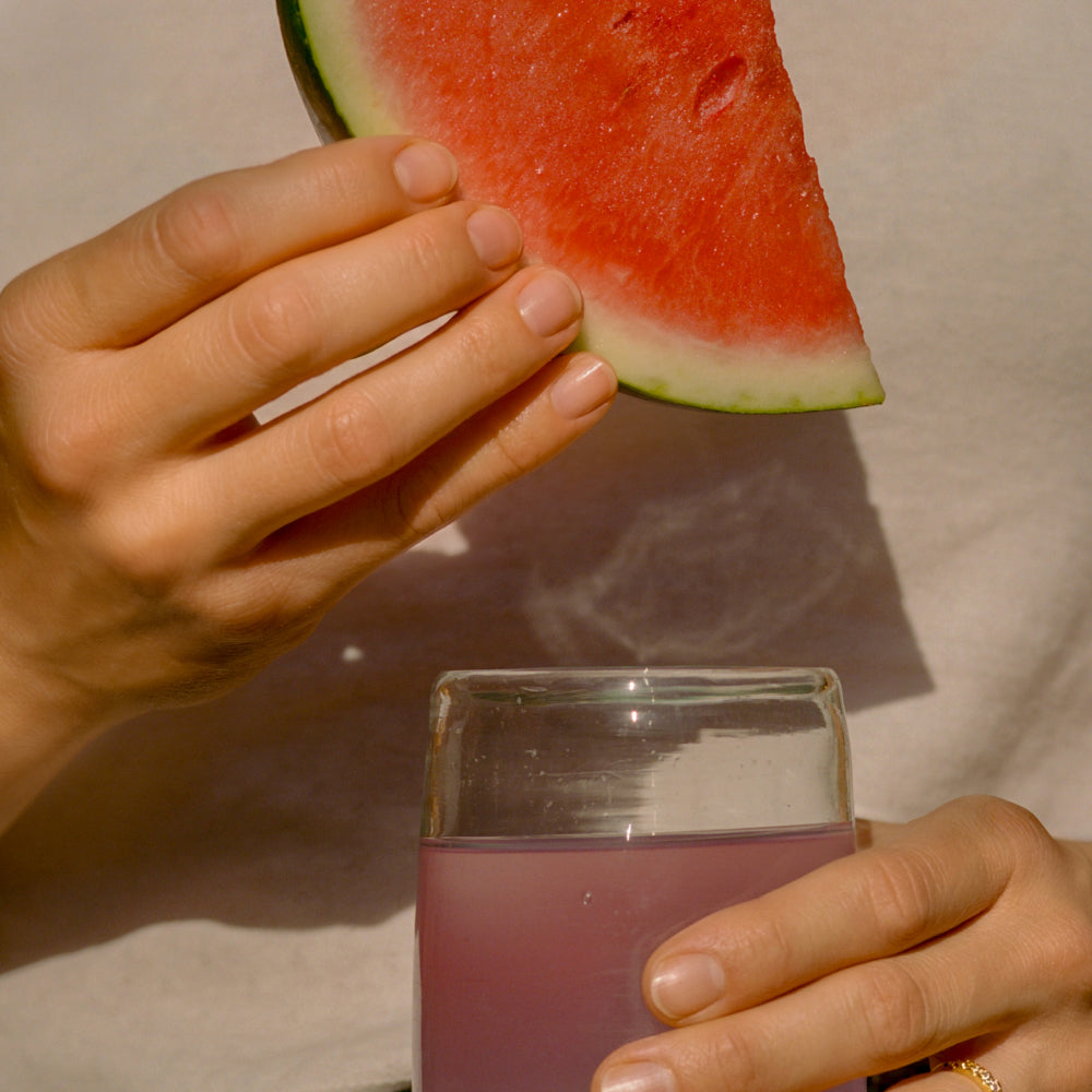 A close-up lifestyle shot of a person holding a glass of pinkish-purple juice in one hand and a slice of watermelon in the other, set against a neutral beige background.