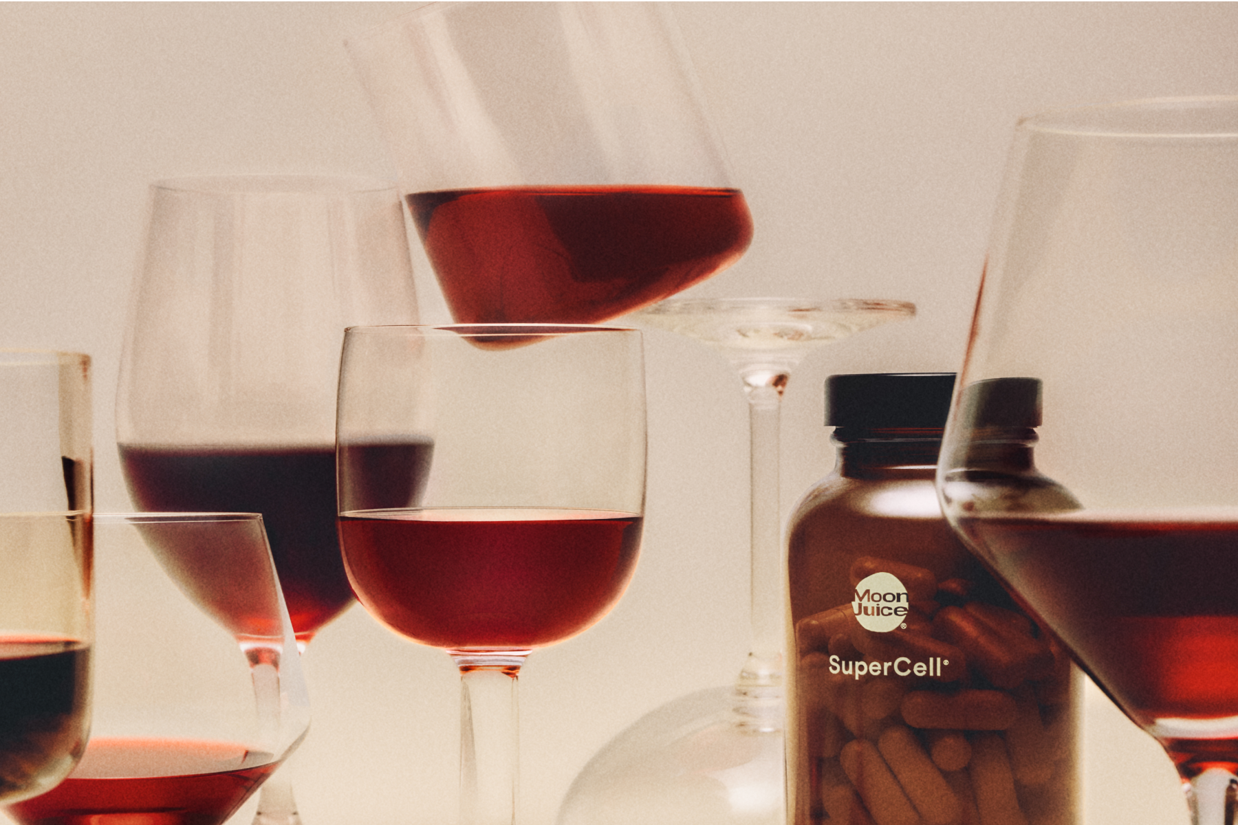 A bottle labeled SuperCell surrounded by several wine glasses filled with red wine, photographed in warm, dim lighting against a soft beige background.
