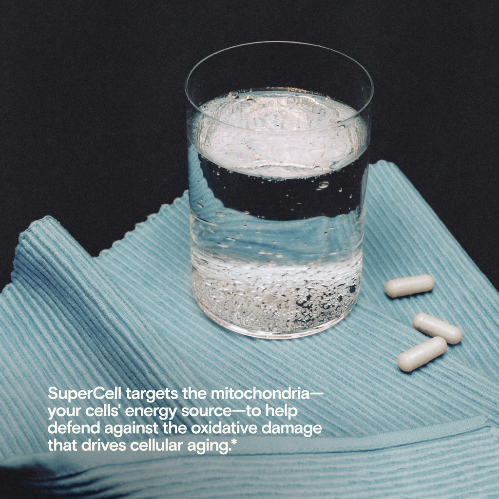 A clear glass cup filled with water sits on a textured light blue cloth against a dark background, with three white capsules placed beside it. White text at the bottom reads, 'SuperCell targets the mitochondria—your cells' energy source—to help defend against the oxidative damage that drives cellular aging.