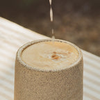 A close-up of a speckled beige ceramic tumbler filled with a light tan beverage, with a thin stream of liquid dripping into the surface. The cup sits on a white slatted surface with a blurred dark background.
